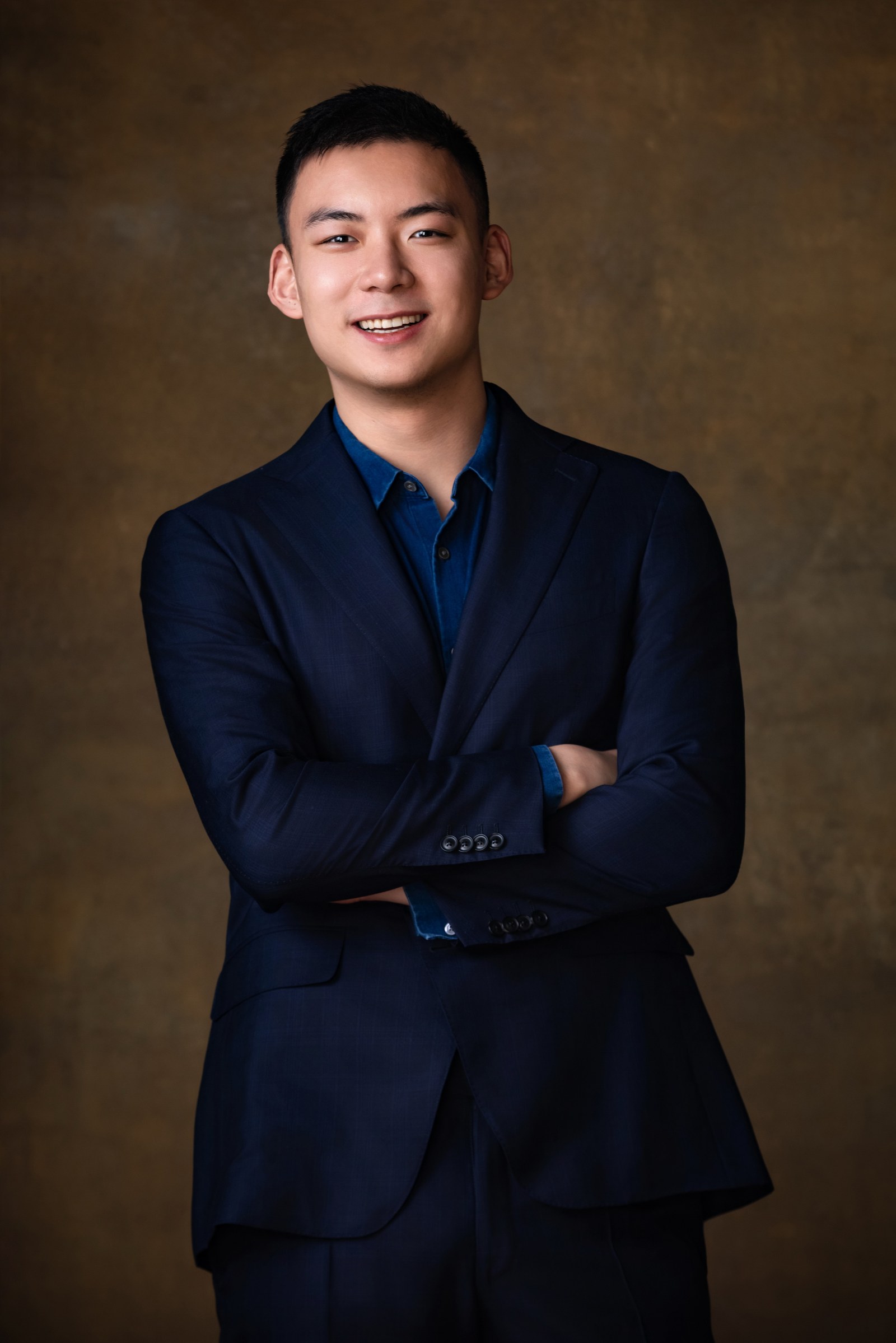 Tony Shen, smiling with arms crossed, wearing a navy suit jacket over a blue shirt against a warm brown studio backdrop.
